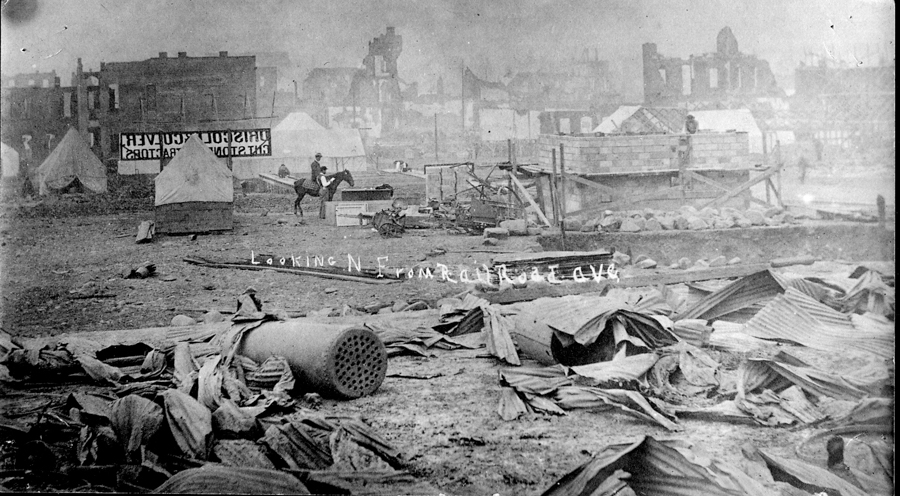 A scene of destruction with debris scattered across the ground. In the background are several tents, one with a sign that reads "U.S. GOVT. CLOTHING." A man on horseback is visible near some wooden structures and piles of rocks. The background shows the ruins of buildings. The text "Looking N. From R. Road Ave" is visible in the foreground.