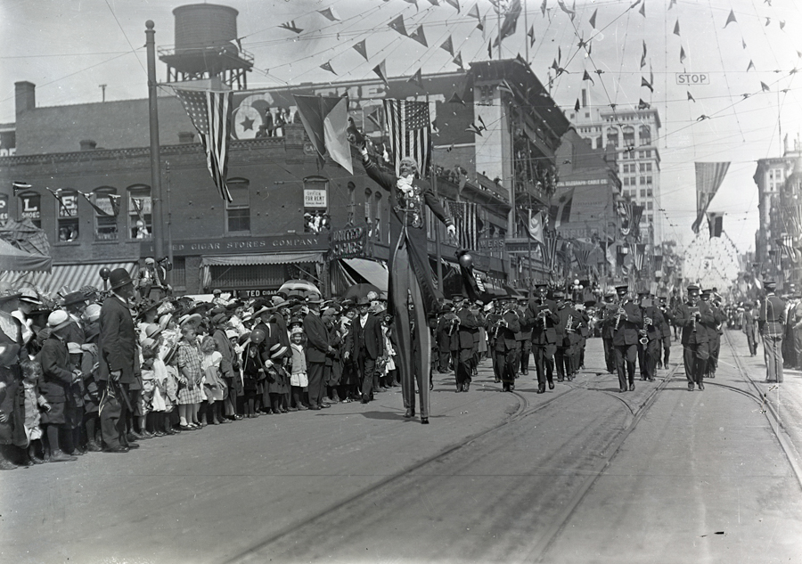 A parade scene on a city street with a crowd of people gathered on the left, including children and adults. Banners and flags are strung overhead. A person on stilts is visible in the middle, and a marching band with instruments like saxophones and trumpets can be seen coming down the street. Buildings line both sides of the street, with some signs visible, including "TED Cigar Stores Company." A "STOP" sign is hanging high above the street.