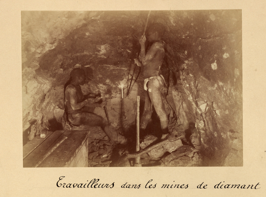 Two men working inside a mine. One man is sitting and appears to be examining something in his hands, while the other stands and holds a tool. The surrounding walls are rocky. The text at the bottom reads, "Travailleurs dans les mines de diamant."