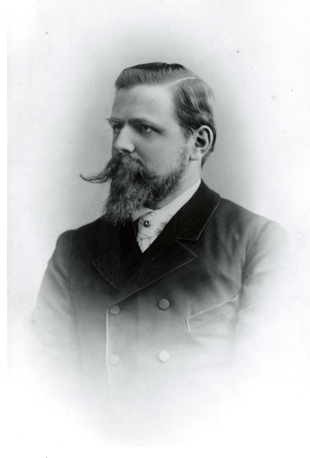 A man with a mustache and beard is wearing a double-breasted coat and a high-collared shirt, looking to the side.