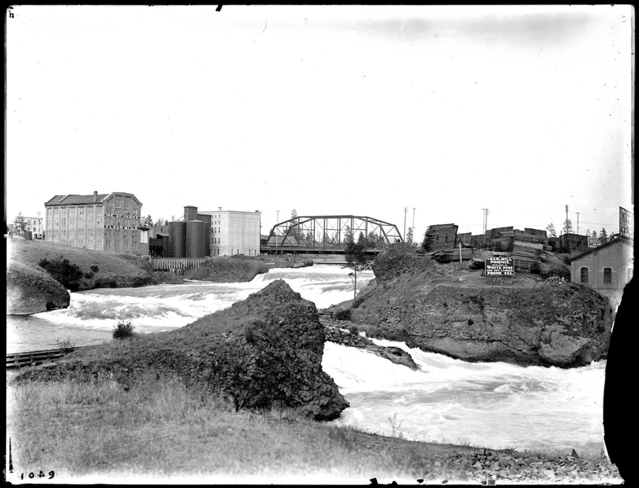 A view of a river with strong rapids, flowing past a rocky landscape. On the left, there are several industrial buildings, including a large brick structure with multiple windows. To the right, stacks of lumber are piled on a rocky hill, with a sign nearby that reads "E. A. WILL & CO SHINGLES WHITE PINE PRICES 1.25X." In the background, a metal truss bridge spans the river, with additional buildings and signs visible in the distance.