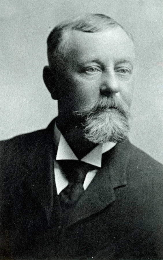 A man with a mustache and a short beard is shown wearing a suit jacket, a white shirt, and a dark tie. He has short, neatly combed hair and is looking slightly to the side.