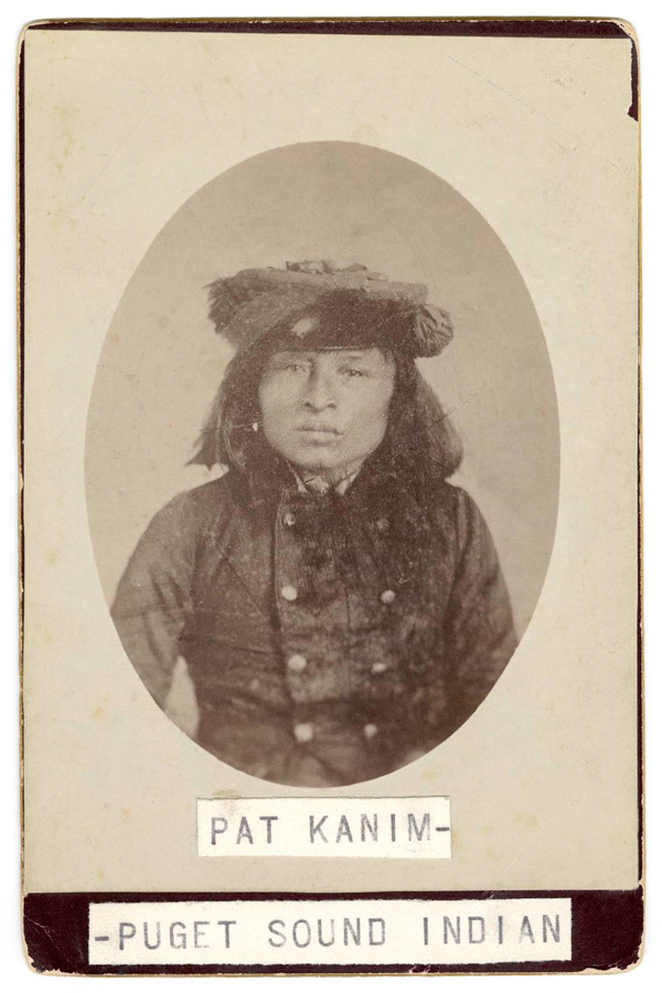 A person wearing a double-breasted coat and a hat adorned with feathers. The text below reads "Pat Kanim - Puget Sound Indian."