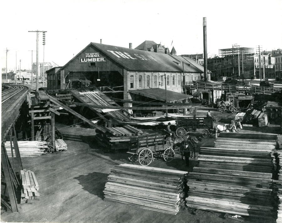 A large industrial site with a building labeled "The Sawmill Phoenix Lumber." Stacks of lumber are arranged throughout the area. Wooden ramps connect the building to other parts of the yard. Horses and wagons are visible, some marked with "Phoenix Lumber Company" and the address "Wholesale, Lumber, Coal, Wood." Railway tracks run along one side, and there are several buildings and smokestacks in the background.