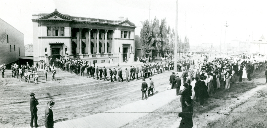 A large group of people gathered outside a two-story building with columns and ornate architecture. Many individuals are in lines, while others form a crowd on the sidewalk. Trees and additional buildings are visible in the background. Several people are riding bicycles or standing with them, and some are dressed in hats and formal clothing.