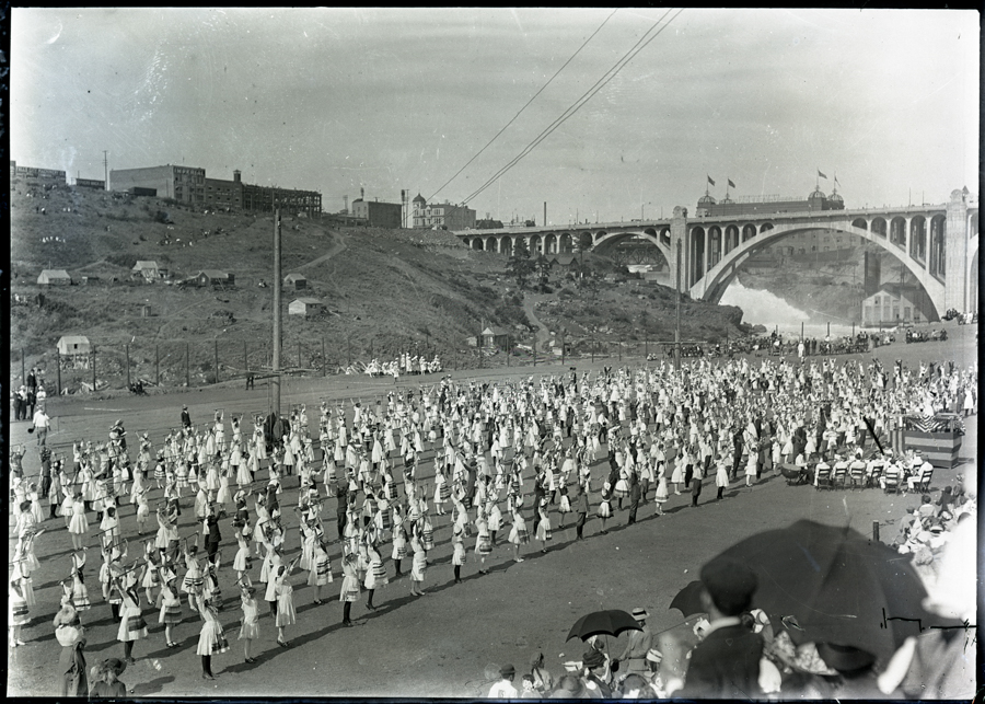 A large group of people are gathered in an open area, dressed in uniform outfits, possibly participating in a coordinated event or parade. In the background, there's a hillside with scattered buildings and a large bridge with arches spanning a river. A crowd of spectators is sitting, some holding umbrellas, watching the event. Flags are visible on the bridge.