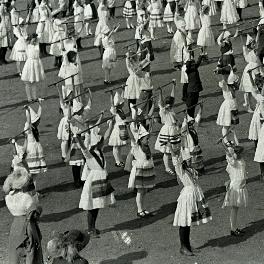 A group of young children dressed in traditional clothing are standing in rows on an open field, each holding a stick above their heads. Some children are wearing pointed hats and their outfits have striped patterns. They appear to be participating in a coordinated activity or performance.