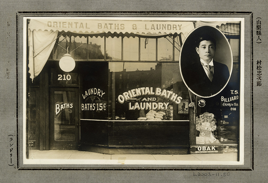 A storefront with a sign reading "Oriental Baths and Laundry" and "Laundry Baths 15¢." The address "210" is above the entrance to one side. There are towels displayed in the window. On the side, there is an oval portrait of a man in a suit. Text on the right side advertises "T.S. Billiard Cigars & Tob. 208" and "OBAK Mouthpiece Cigarettes." Japanese characters are present on the right and left borders.