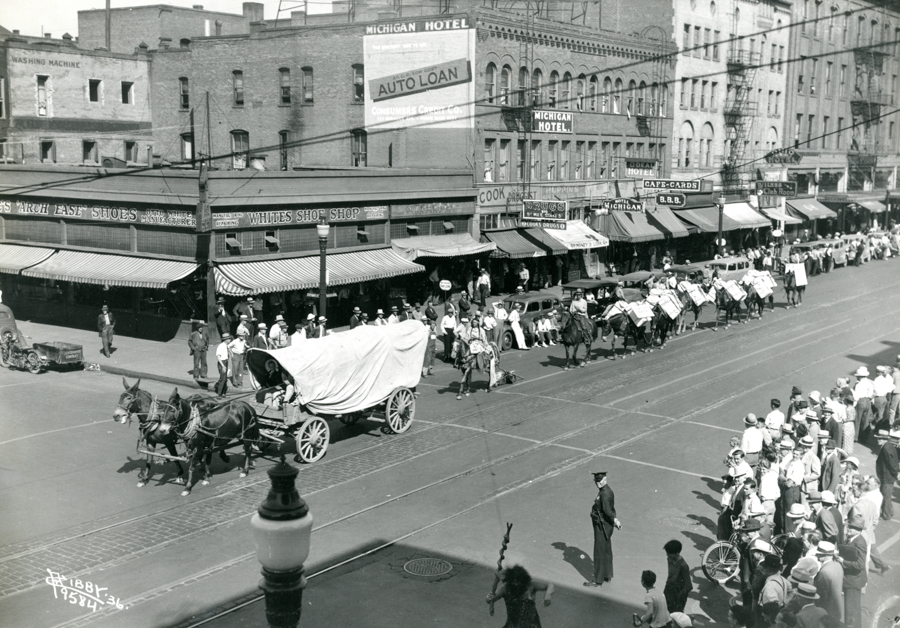 A street scene with a parade featuring covered wagons and people on horseback moving along a city thoroughfare. Onlookers, some wearing hats, are gathered on the sidewalk watching the procession. Nearby buildings have awnings and signage, including "Whites Shoe Shop," "Hub Clothing," "Michigan Hotel," "Auto Loan," and "Drugs." There are several automobiles parked along the street. A man in uniform is standing in the street near the crowd.