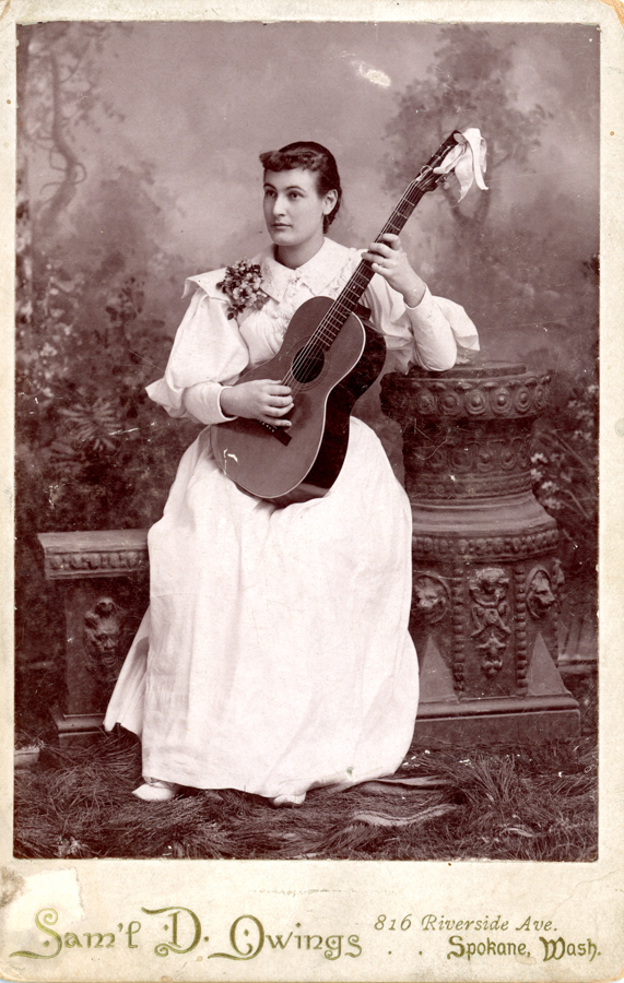 A woman in a white dress holds a guitar while seated between an ornate pedestal and a decorative stone. She has flowers pinned to her dress. The background features a blurred natural scene. Text at the bottom reads "Sam'l D. Owings, 816 Riverside Ave., Spokane, Wash."