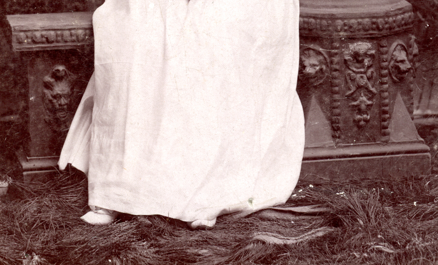 A person wearing a long white garment standing on what appears to be straw or dry grass. Behind them is a stone structure with ornate carvings, including a face and decorative patterns.