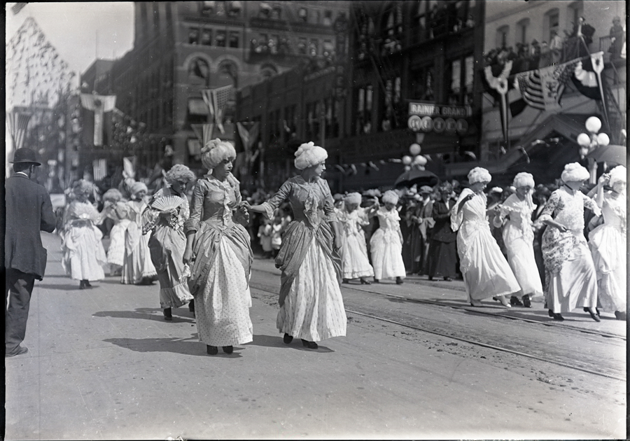 A group of women dressed in elaborate period costumes are parading down a street. They wear long, elegant dresses with large, decorative hats or wigs. Onlookers line the street, and buildings in the background are decorated with flags and banners. A sign in the background reads "RAINIER GRAND HOTEL."