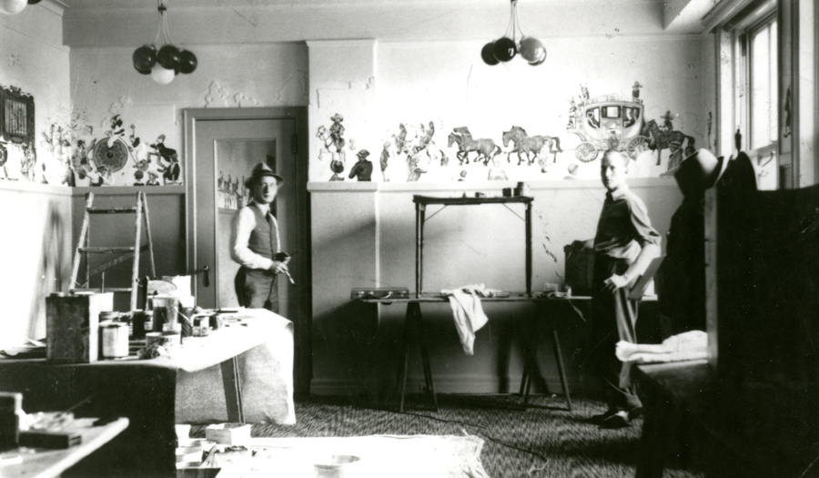 Two men in a room with painted wall decorations showing horses and a carriage. One man is wearing a hat and holding a brush, while the other stands near a table. The room contains a ladder and various paint supplies.