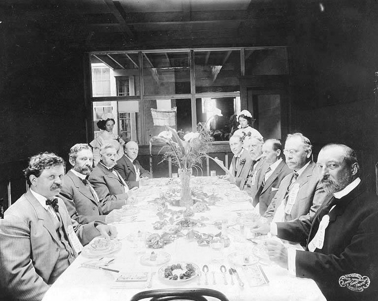 A group of men wearing suits and ties are seated on either side of a long dining table, set with plates, cutlery, and floral arrangements in the center. Two women stand in the background by a window. A sign in the lower right corner reads "C. M. Hayes Pittsburgh, PA."