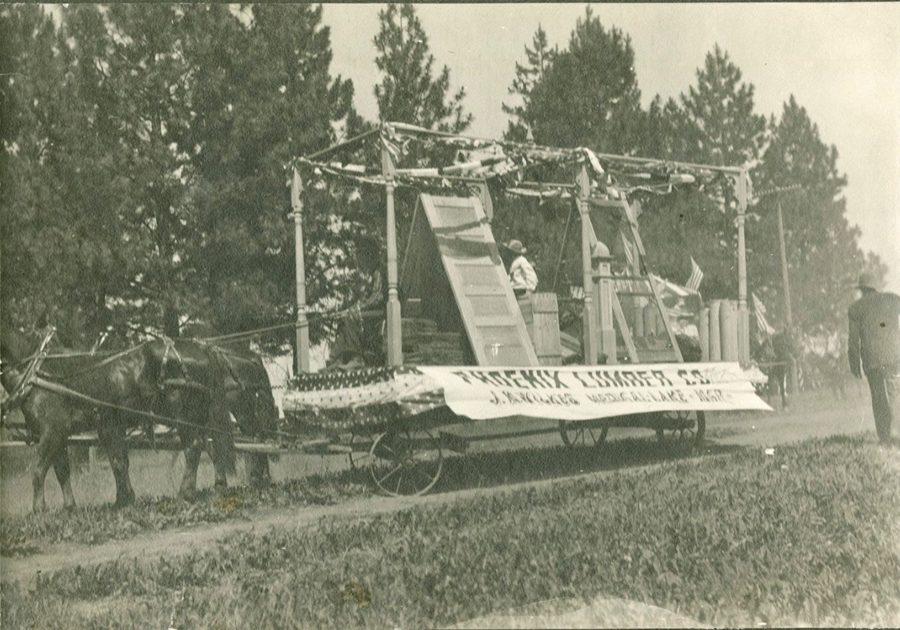 A float in a parade pulled by horses. It is decorated with flags and features wooden panels. A banner on the side reads "Phoenix Lumber Co. J. Bendickson, Mgr. Lake Mary." Trees and a person walking are in the background.