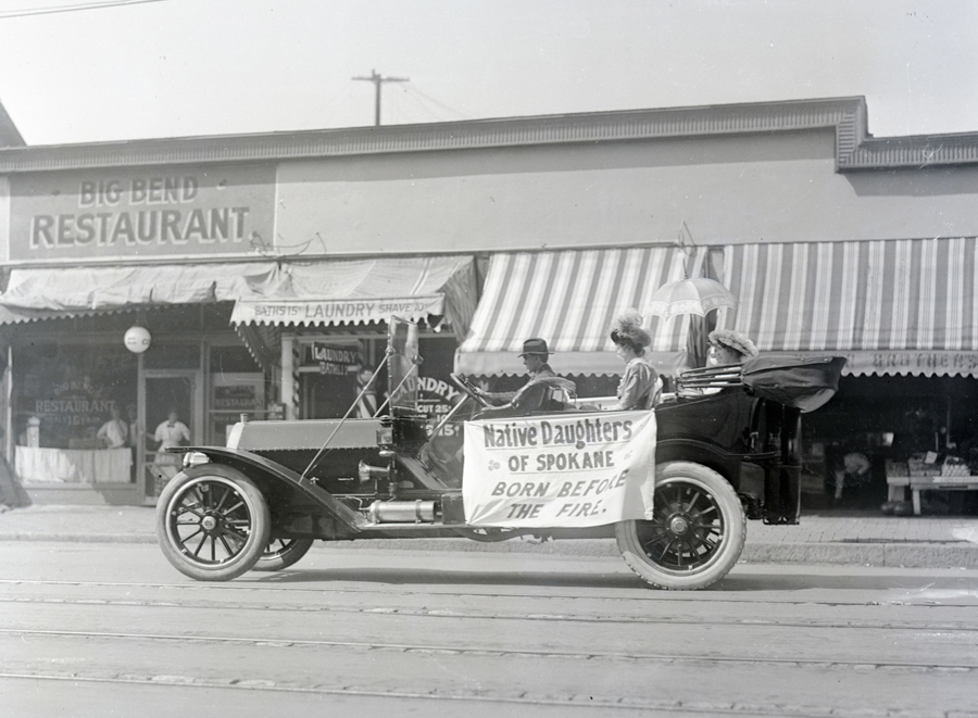 A vintage car is parked on a street, with a sign on the side reading "Native Daughters of Spokane, Born Before the Fire." Two women in hats sit in the back of the car, one holding an umbrella, while a man in the front appears to be driving. In the background is a building with a sign that says "Big Bend Restaurant" and another advertising a laundry service. The awnings over the windows have stripes.