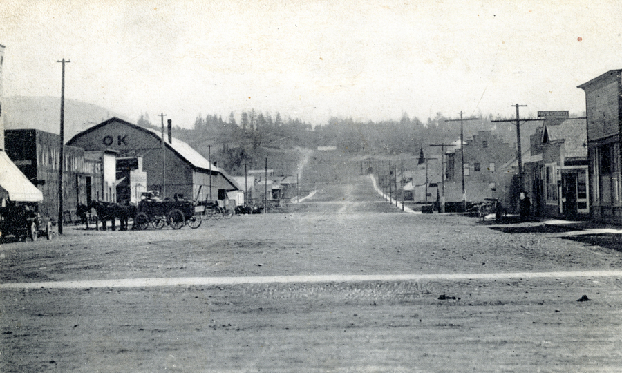 A wide street lined with wooden buildings stretches into the distance. On the left, a building with "OK" written on it stands beside a horse-drawn carriage. Utility poles and lampposts are visible along the sidewalks. In the background, there are trees and hills.