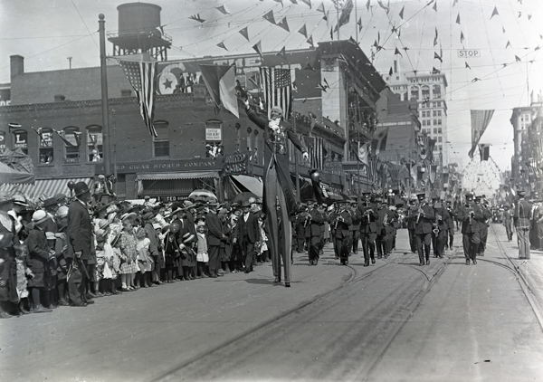 A parade scene on a city street with a crowd of people gathered on the left, including children and adults. Banners and flags are strung overhead. A person on stilts is visible in the middle, and a marching band with instruments like saxophones and trumpets can be seen coming down the street. Buildings line both sides of the street, with some signs visible, including 