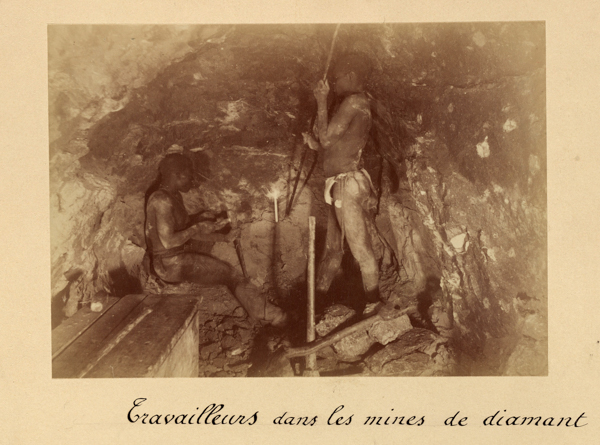 Two men working inside a mine. One man is sitting and appears to be examining something in his hands, while the other stands and holds a tool. The surrounding walls are rocky. The text at the bottom reads, 