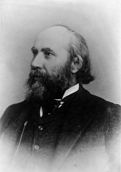 A bearded man with a receding hairline and wearing a suit is seen in profile. His attire includes a vest, a jacket, and a tie. The background is plain.