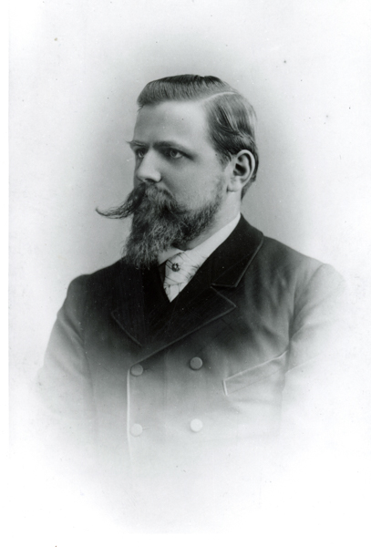 A man with a mustache and beard is wearing a double-breasted coat and a high-collared shirt, looking to the side.