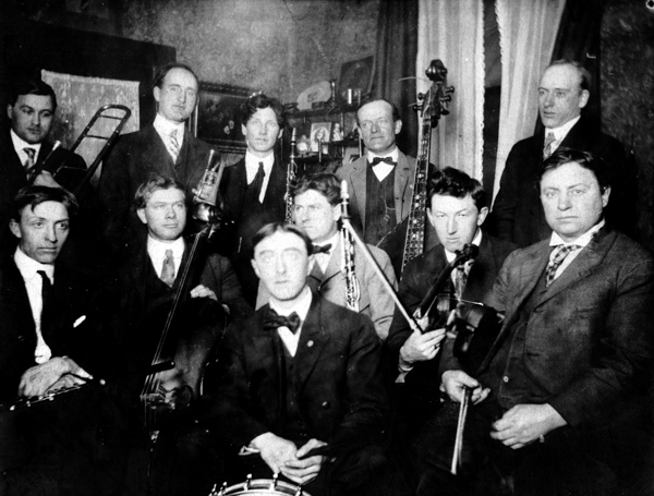 A group of eleven men sit and stand closely together in a room, holding various musical instruments such as trombones, violins, a clarinet, a trumpet, and a drum. Most are dressed in suits with bow ties or ties. A curtain and wall decorations are visible in the background.