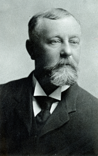 A man with a mustache and a short beard is shown wearing a suit jacket, a white shirt, and a dark tie. He has short, neatly combed hair and is looking slightly to the side.