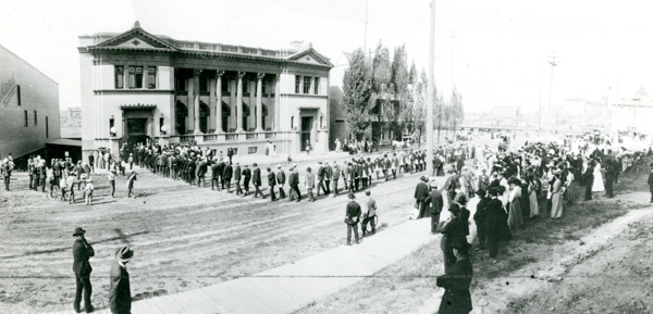 A large group of people gathered outside a two-story building with columns and ornate architecture. Many individuals are in lines, while others form a crowd on the sidewalk. Trees and additional buildings are visible in the background. Several people are riding bicycles or standing with them, and some are dressed in hats and formal clothing.