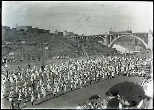 A large group of people are gathered in an open area, dressed in uniform outfits, possibly participating in a coordinated event or parade. In the background, there's a hillside with scattered buildings and a large bridge with arches spanning a river. A crowd of spectators is sitting, some holding umbrellas, watching the event. Flags are visible on the bridge.