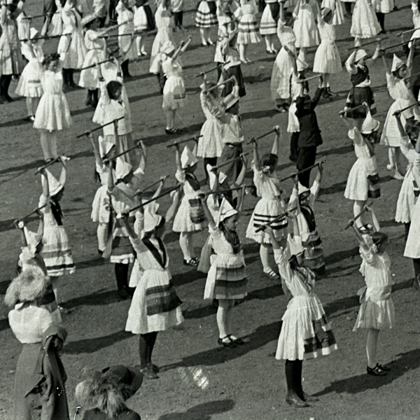 A group of young children dressed in traditional clothing are standing in rows on an open field, each holding a stick above their heads. Some children are wearing pointed hats and their outfits have striped patterns. They appear to be participating in a coordinated activity or performance.