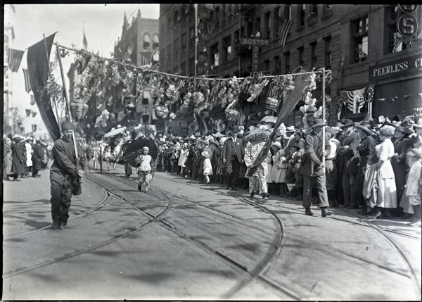 A street scene with a parade featuring people carrying banners and lanterns decorated with flowers. A child holding an umbrella walks in the middle of the street. Both sides of the street are lined with spectators, many wearing hats. Buildings in the background have signs reading 