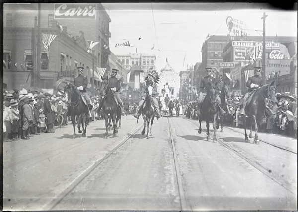 A parade on a busy street with five people riding horses. Four are in uniform, and one is wearing a headdress. Crowds line both sides of the street, with flags and banners hanging from surrounding buildings. Signs in the background include 