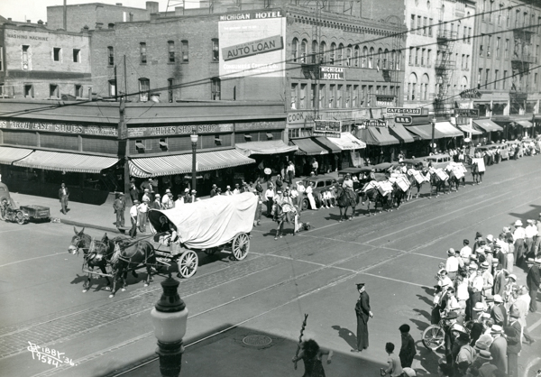 A street scene with a parade featuring covered wagons and people on horseback moving along a city thoroughfare. Onlookers, some wearing hats, are gathered on the sidewalk watching the procession. Nearby buildings have awnings and signage, including 