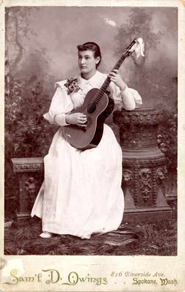 A woman in a white dress holds a guitar while seated between an ornate pedestal and a decorative stone. She has flowers pinned to her dress. The background features a blurred natural scene. Text at the bottom reads 