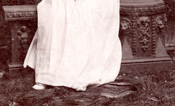 A person wearing a long white garment standing on what appears to be straw or dry grass. Behind them is a stone structure with ornate carvings, including a face and decorative patterns.