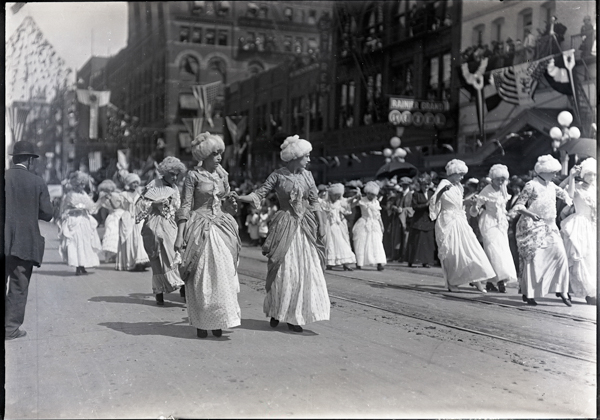 A group of women dressed in elaborate period costumes are parading down a street. They wear long, elegant dresses with large, decorative hats or wigs. Onlookers line the street, and buildings in the background are decorated with flags and banners. A sign in the background reads 