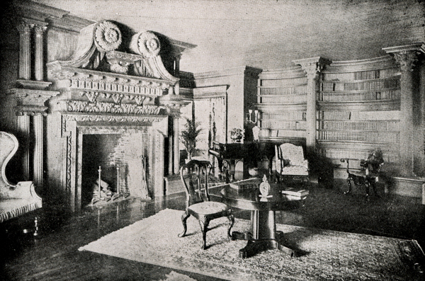 An ornate room features a large, intricately carved fireplace with two decorative columns on either side. To the right, there is a curved bookshelf filled with books. A round table with a small decorative item and an open book is positioned at the center of the room, surrounded by elegant chairs. In the background, a potted plant and table with a lamp are visible, along with a curved sofa and additional chairs.