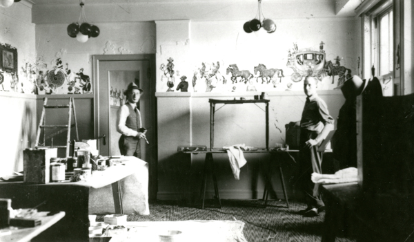 Two men in a room with painted wall decorations showing horses and a carriage. One man is wearing a hat and holding a brush, while the other stands near a table. The room contains a ladder and various paint supplies.