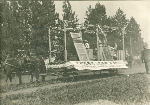 A float in a parade pulled by horses. It is decorated with flags and features wooden panels. A banner on the side reads 