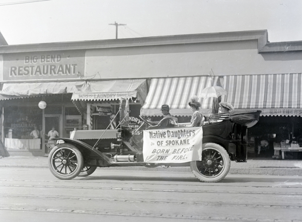 A vintage car is parked on a street, with a sign on the side reading 