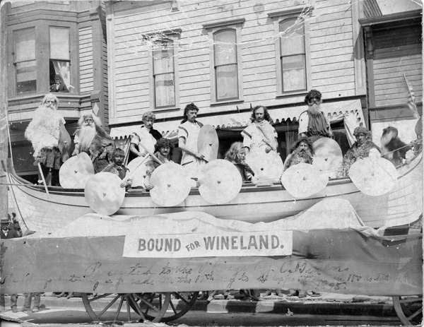 A group of people dressed in costumes stand on a float shaped like a boat with circular shields. The float has a sign that reads 