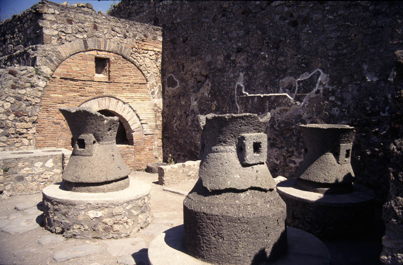 This image shows the ruins of an ancient bakery. There is a large brick oven in the back that forms an entire wall of the bakery. There are three large stone mills with supports to attach the contraptions that would have turned the donkey's movement into the power that would turn the stones to grind flour.